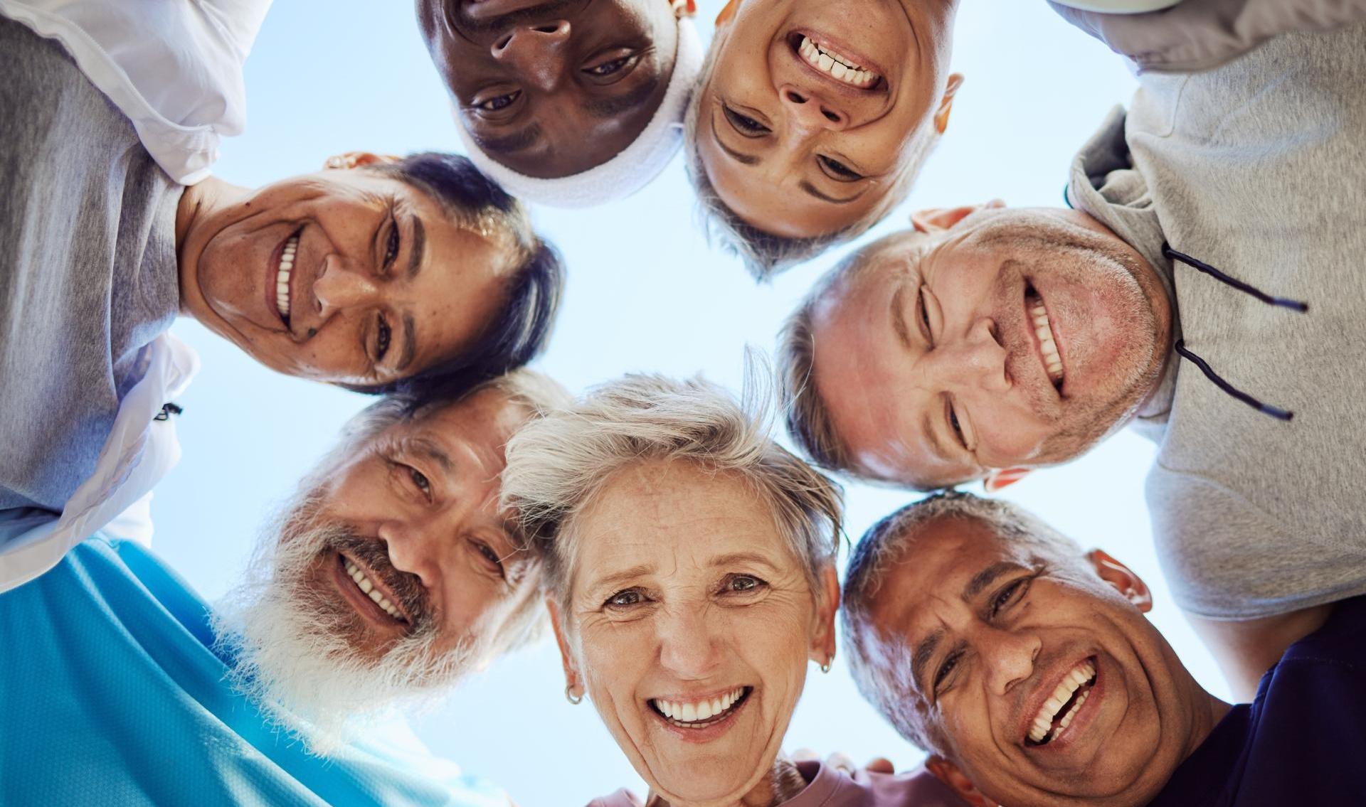 Seven adults arranged in a close circular connecting  huddle, faces pointing inward toward the camera expressing joy.