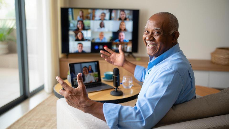 A cheerful older man participates in a video conference from his home office, showcasing a laptop and microphone, with a group of people visible on the screen.