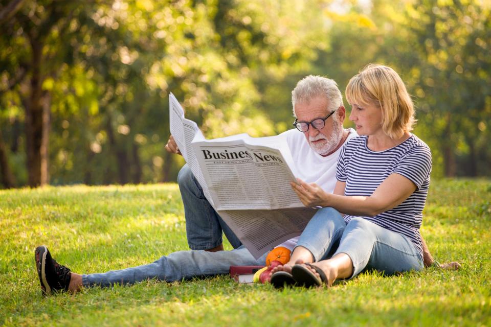 Couple reading the same newspaper while sitting on the grass in a park.