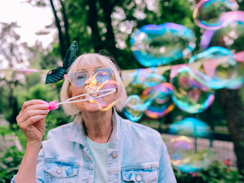 stylish woman with gray hair and in blue glasses and jeans jacket blowing bubbles outdoors.